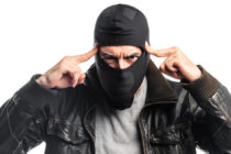 criminoso mascarado apontando com as duas mãos para a própria cabeça, indicando que ele está pensando ou pedindo para alguém pensar