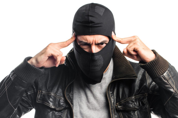 criminoso mascarado apontando com as duas mãos para a própria cabeça, indicando que ele está pensando ou pedindo para alguém pensar